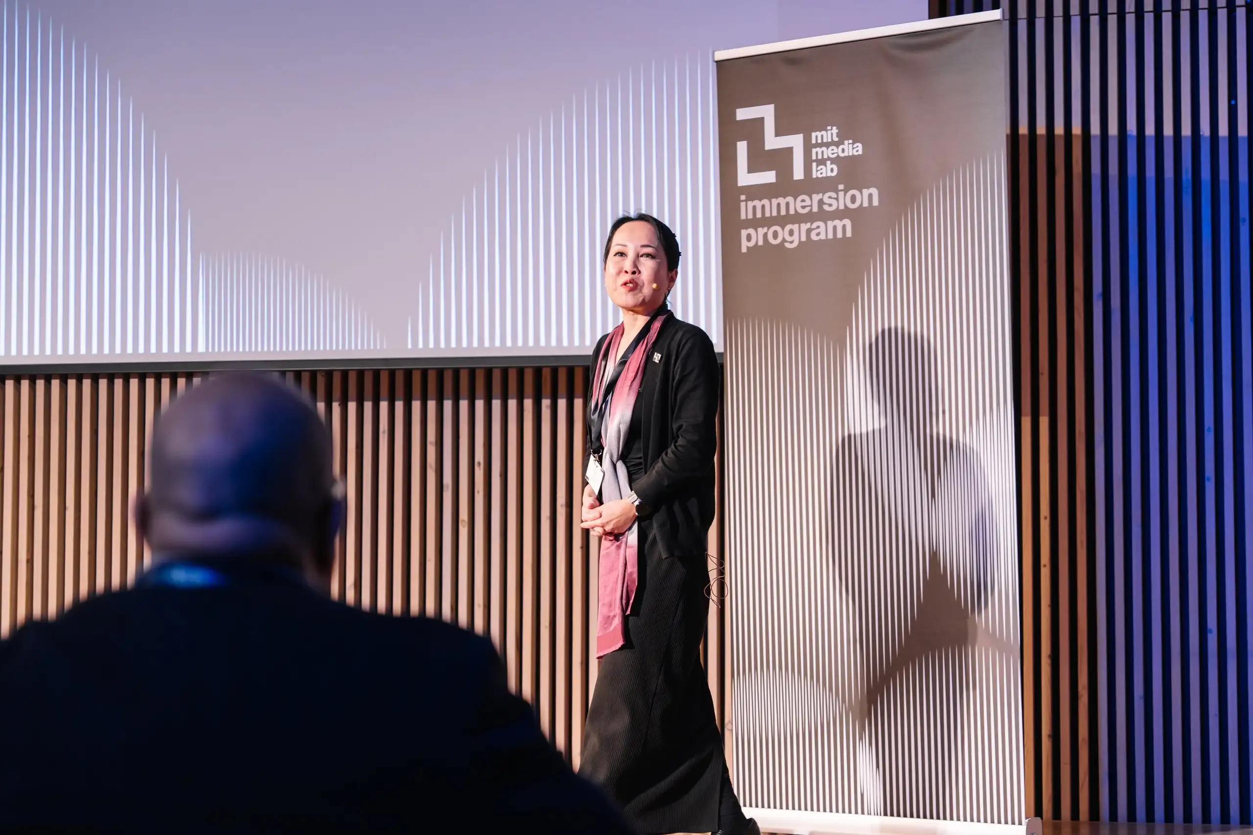 Una ponent parla a l’escenari, al costat del cartell de l’Immersion Program del MIT Media Lab
