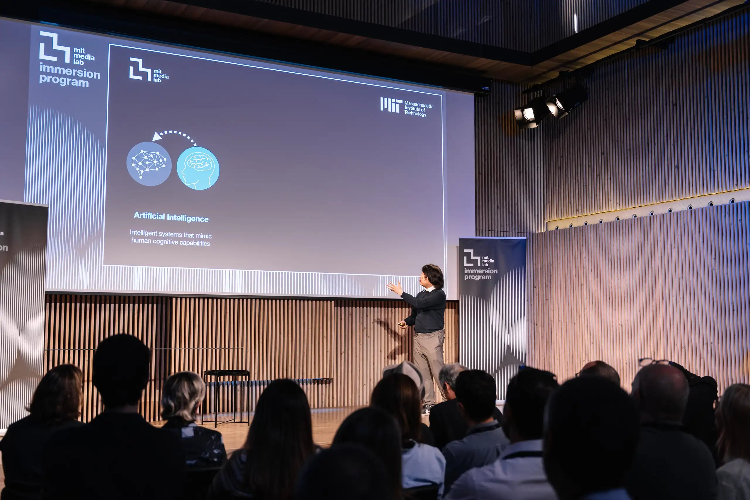 El públic segueix una xerrada sobre intel·ligència artificial projectada a la pantalla de l’escenari