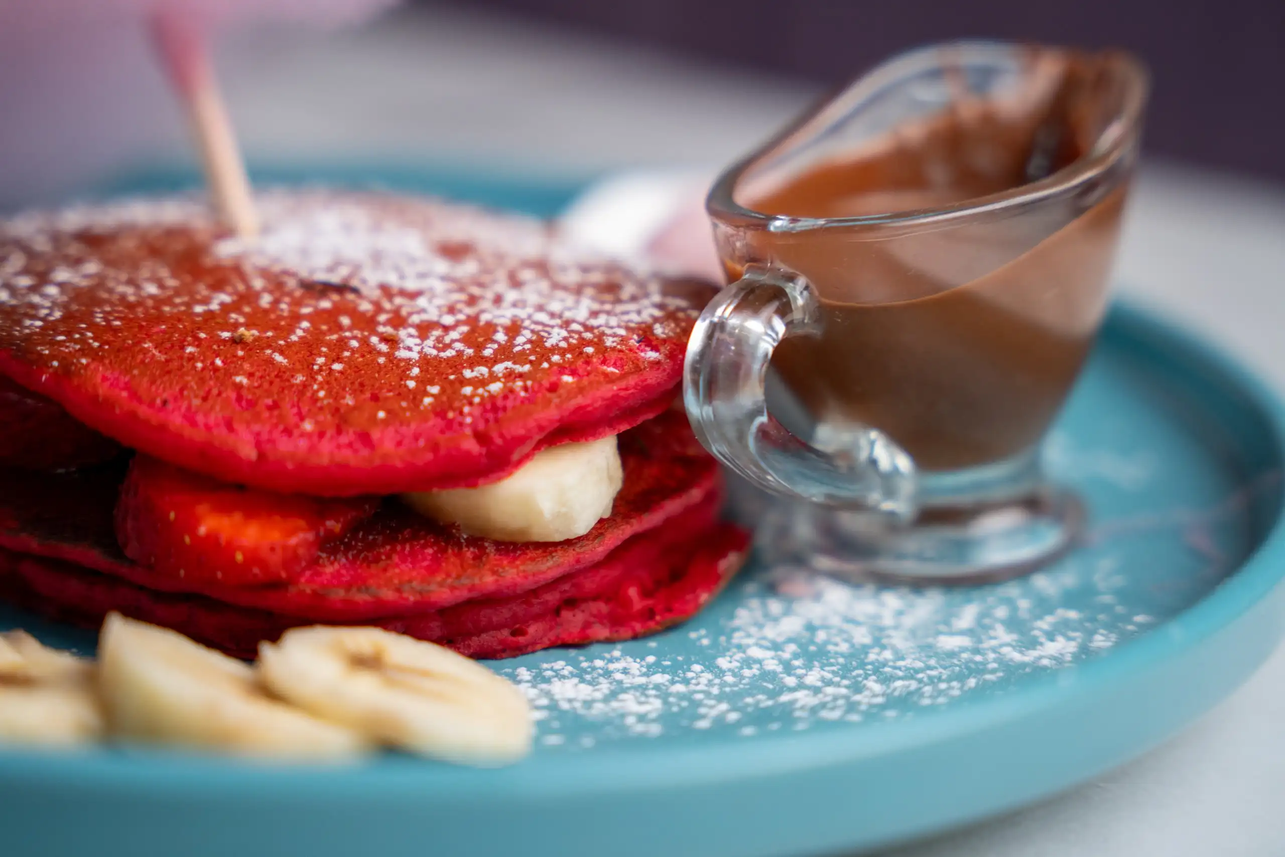 Pancakes vermells amb plàtan, maduixes i sucre glaçat, amb una gerreta de xocolata, en un plat blau