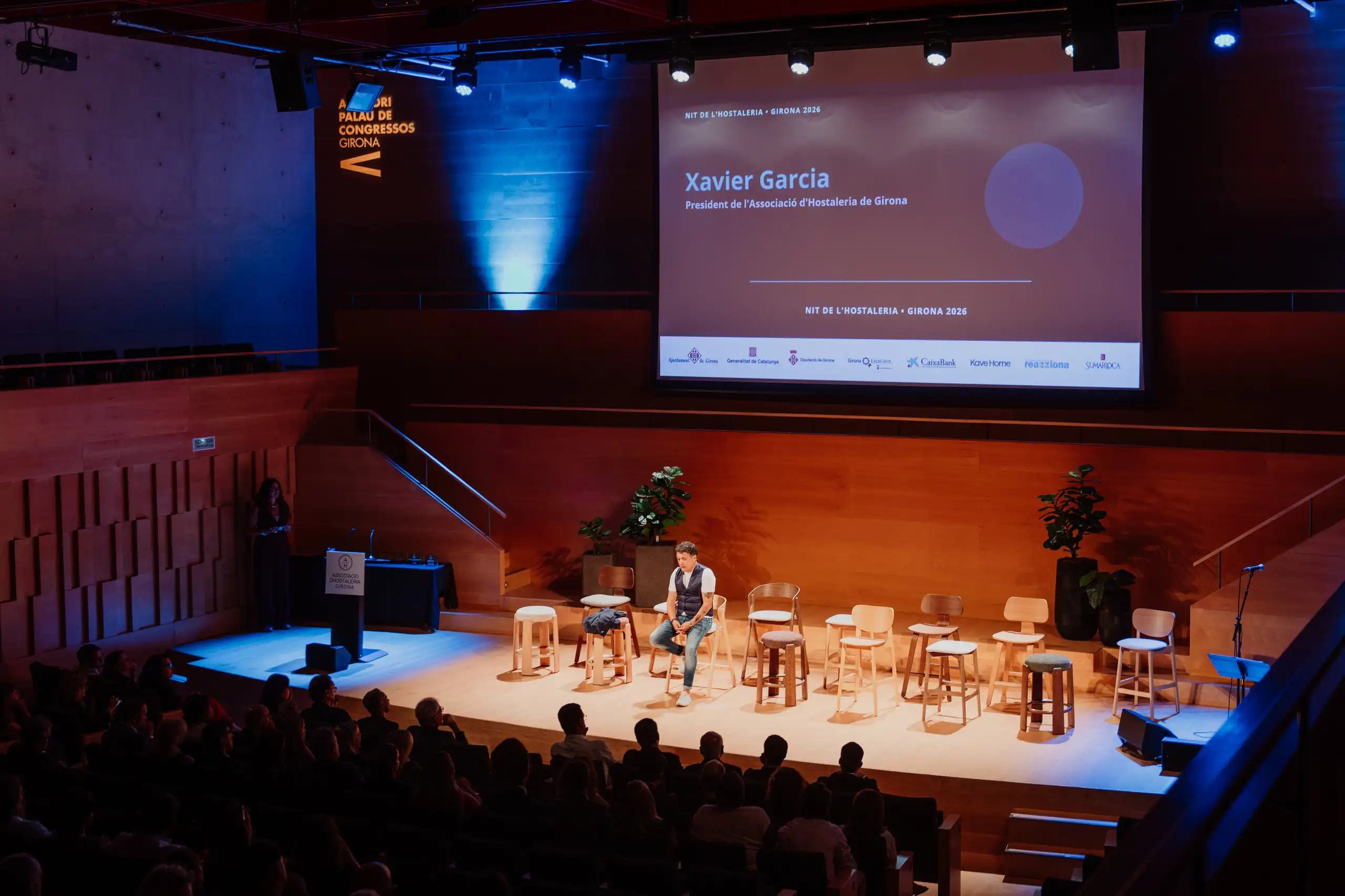 Vista general de l’auditori durant la Nit de l’Hostaleria, amb l’escenari il·luminat en blau i taronja