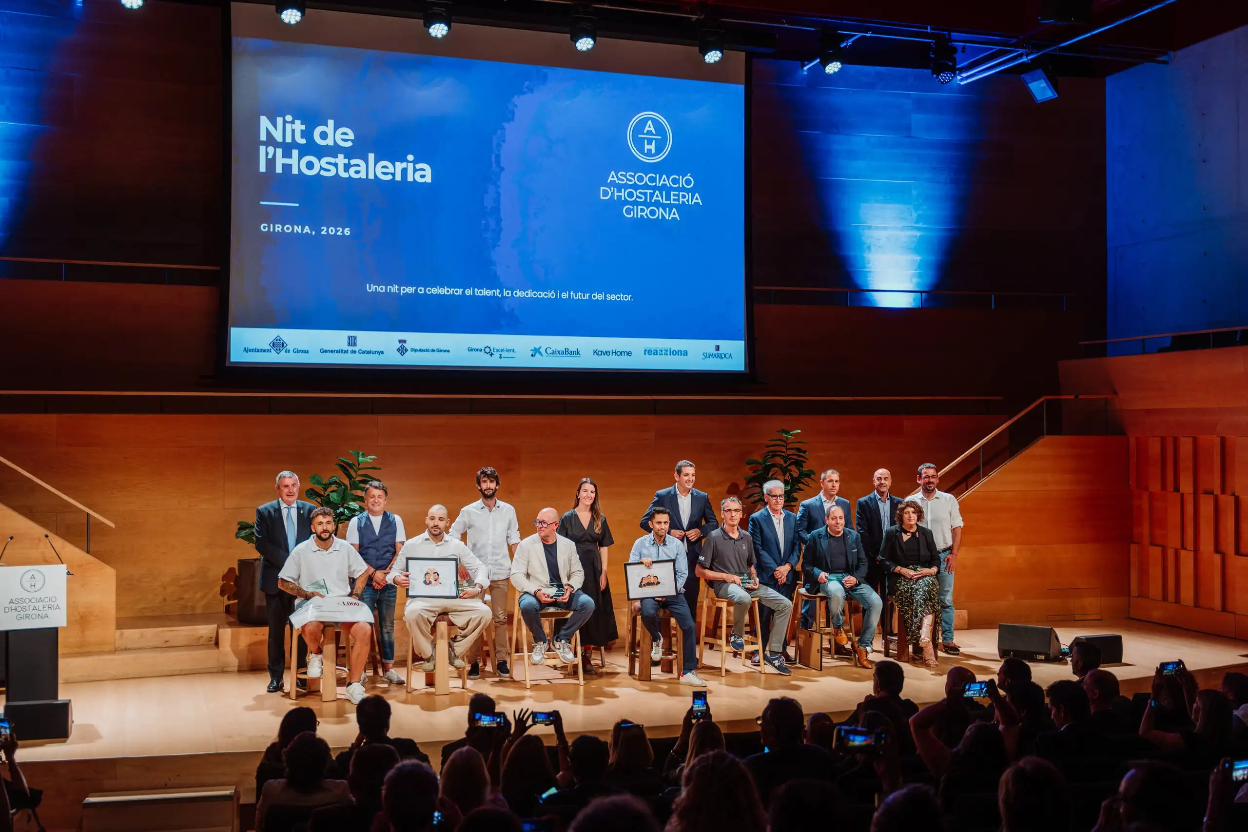 Nombrós grup assegut en fila a l’escenari, davant el públic i la pantalla de la gala