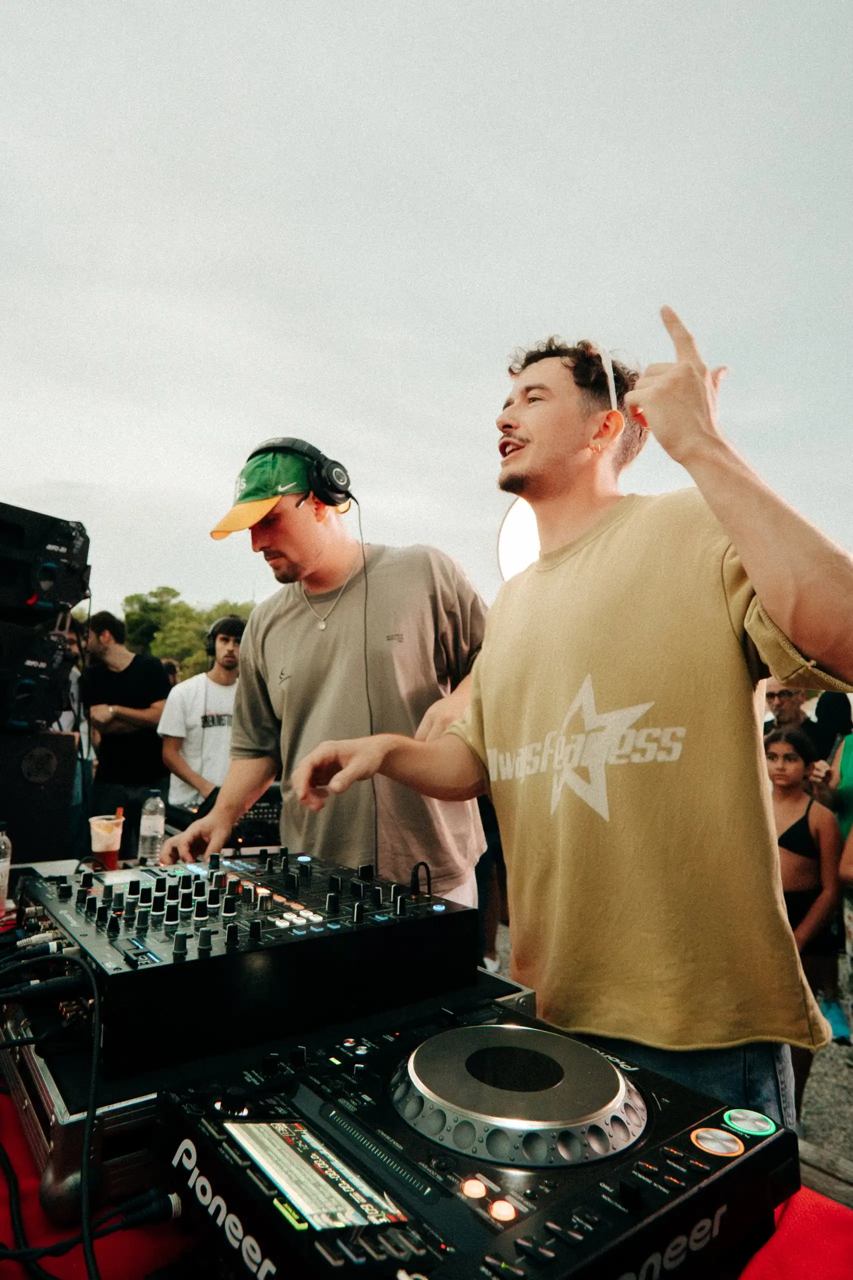 Dos DJ davant la taula de mescles a l’aire lliure, un d’ells assenyalant el cel