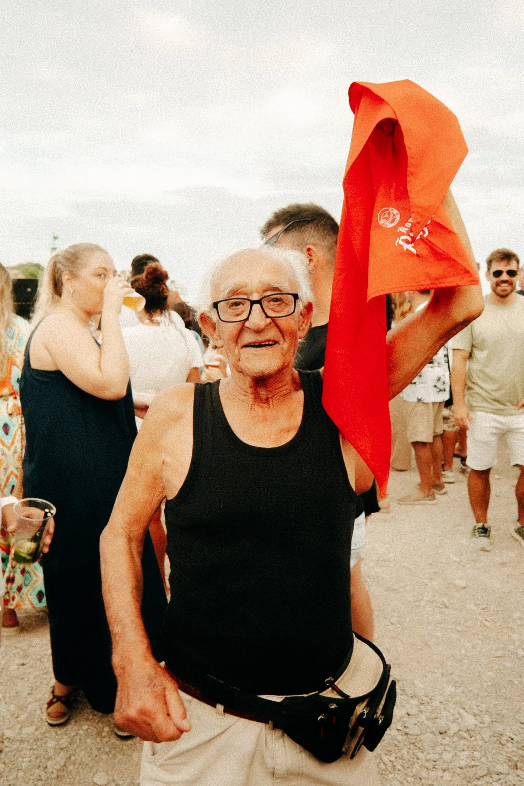 Un senyor gran somriu a càmera amb un mocador vermell a l’espatlla, enmig del públic